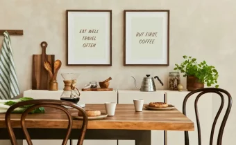 Útulná jídelna s dřevěným stolem, nad nímž visí obrazy s nápisy "EAT WELL TRAVEL OFTEN" a "BUT FIRST, COFFEE", což vytváří vřelou atmosféru.