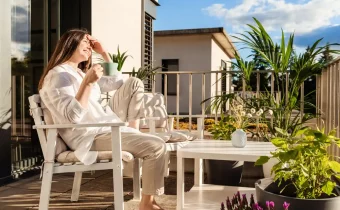 Žena relaxuje na balkóne paneláku s bielym nábytkom a obklopená zelenými rastlinami.
