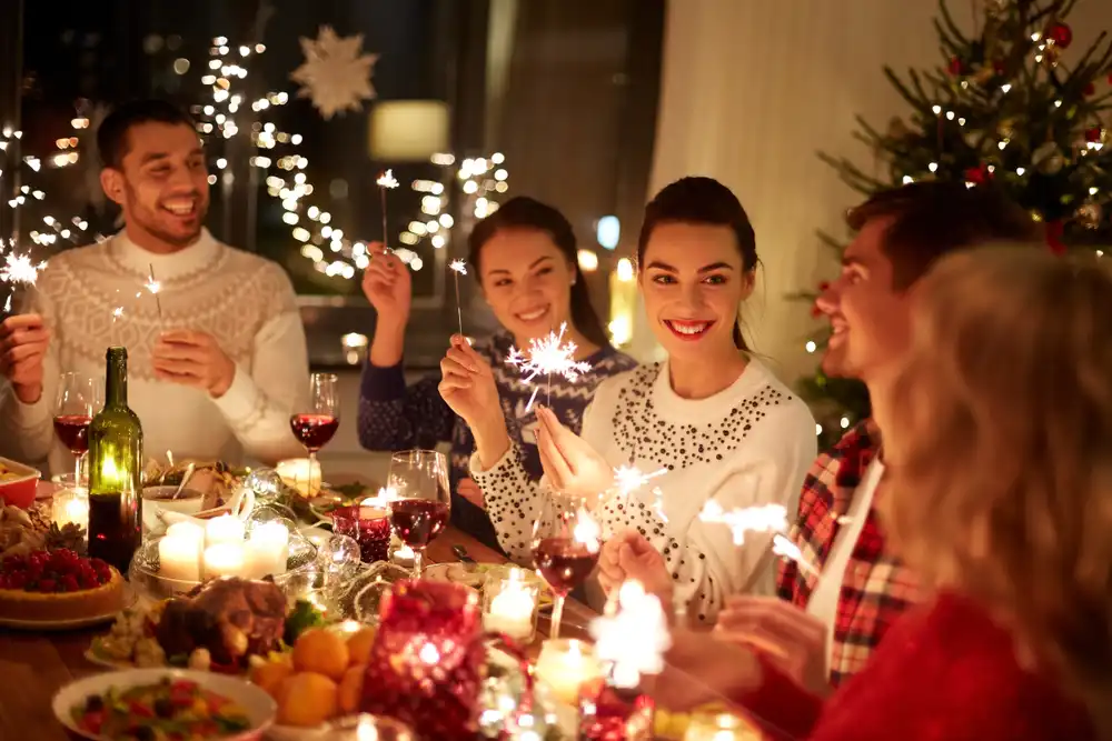 Skupina priateľov si užíva vianočný večierok pri stole osvetlenom sviečkami a vianočnými svetielkami, držia bengálske ohně, pripíjajú a majú na sebe sviatočné svetre, čo vytvára radostnú a priateľskú atmosféru.