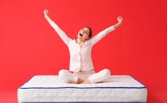 Žena sediaca na matraci vyzerá šťastne a rozpažuje na červenom pozadí, čo symbolizuje dobrý spánok a pohodlie, ktoré kvalitná matrace poskytuje.