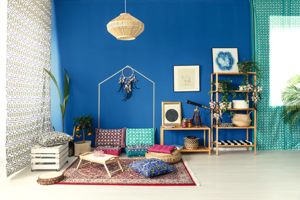 Interiér v boho štýle s výraznou modrou stenou, vzorovanými textíliami, ratanovým lustrom a dekoratívnymi prvkami ako sú vankúše, rastliny a obrazy.