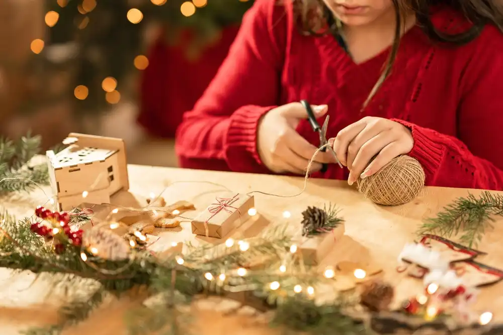 Dievča v červenom svetri tvorí vianočné dekorácie, strihá šnúrku a má pred sebou vianočné svetielka a výzdobu, čo naznačuje tvorbu DIY vianočných ozdôb.