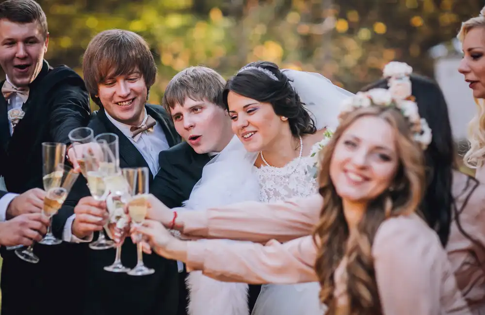 Skupina ľudí oslavujúca na svadbe, držia si poháre so šampanským a pripíjajú, v popredí je družička s kvetinovým vencom na hlave a hostia v elegantných odevách.