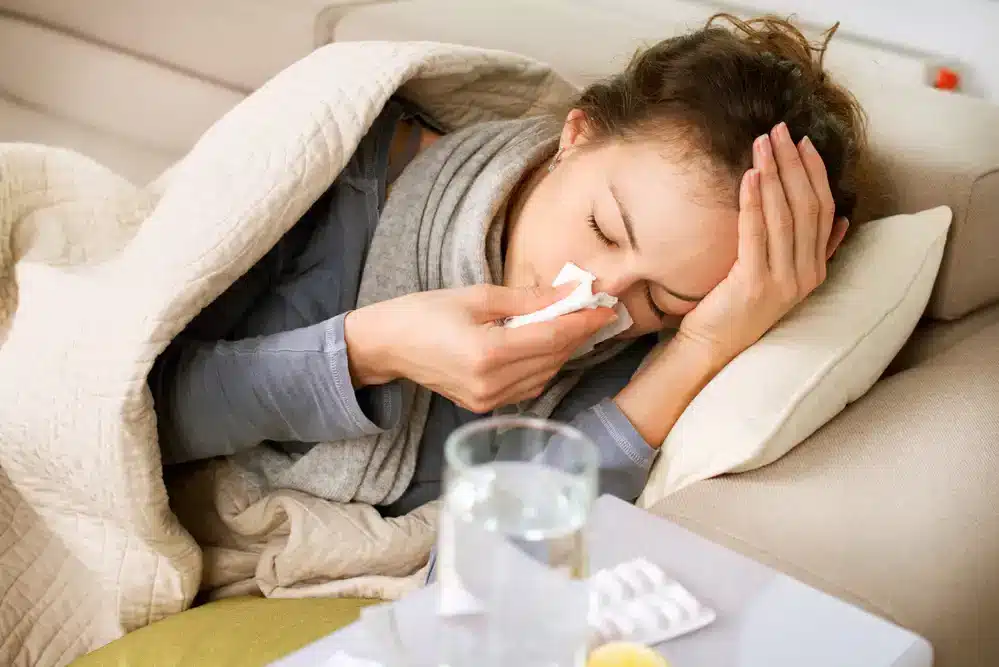 Žena ležiaca na gauči s horúčkou, utiera si nos papierovým vreckovkom, obklopená liekmi a pohárom vody, vyzerá unavene a chorľavo.