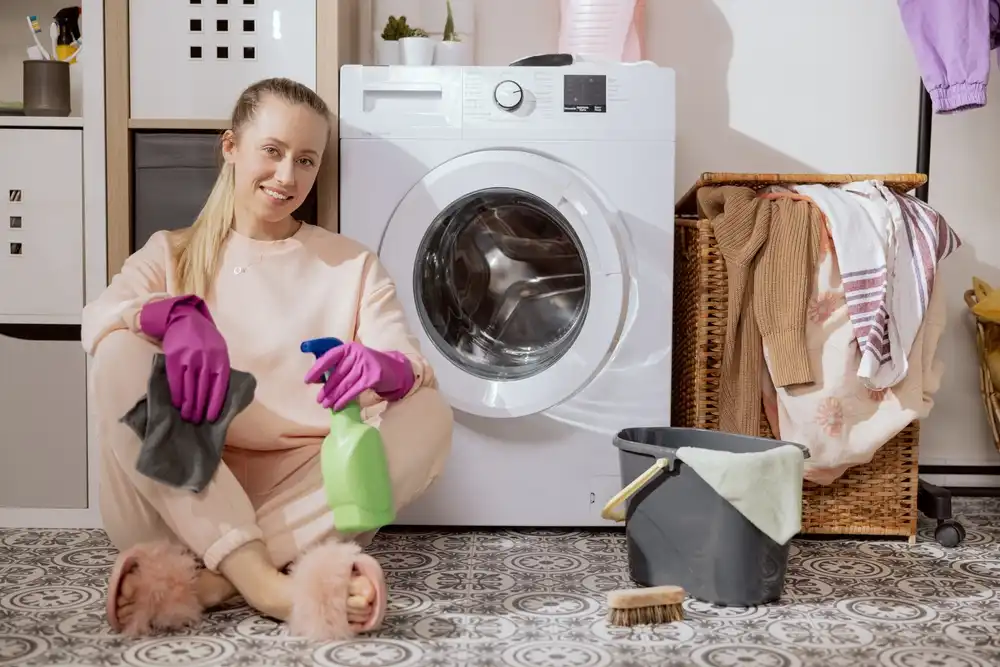 Usmievavá žena sedí na podlahe s čistiacimi prostriedkami a fialovými rukavicami pri otvorenej práčke, pripravená na čistenie.