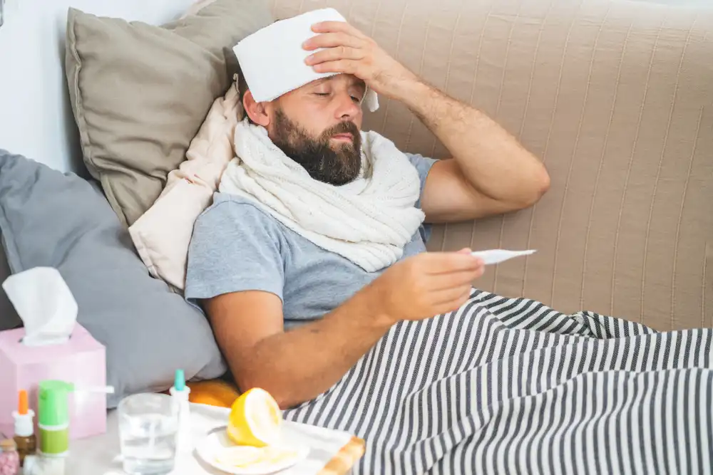 Muž sedí na posteli s uterákom na hlave, teplomerom a liekmi okolo seba, naznačuje horúčku a chorobu.