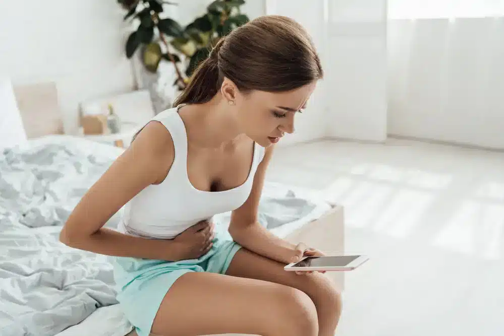 Mladá žena sediaca na posteli, drží sa za brucho a používa tablet, možný znak menštruačných kŕčov alebo vyhľadávania informácií o ich úľave.