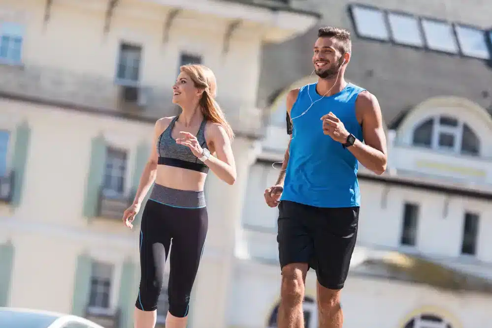 Muž a žena v športovom oblečení bežia vedľa seba na mestskom chodníku. Muž má na sebe modré tričko a čierne šortky, žena nosí sivú športovú podprsenku a čierne legíny. Oboje vyzerajú šťastne a energicky, čo ilustruje radosť z behania.