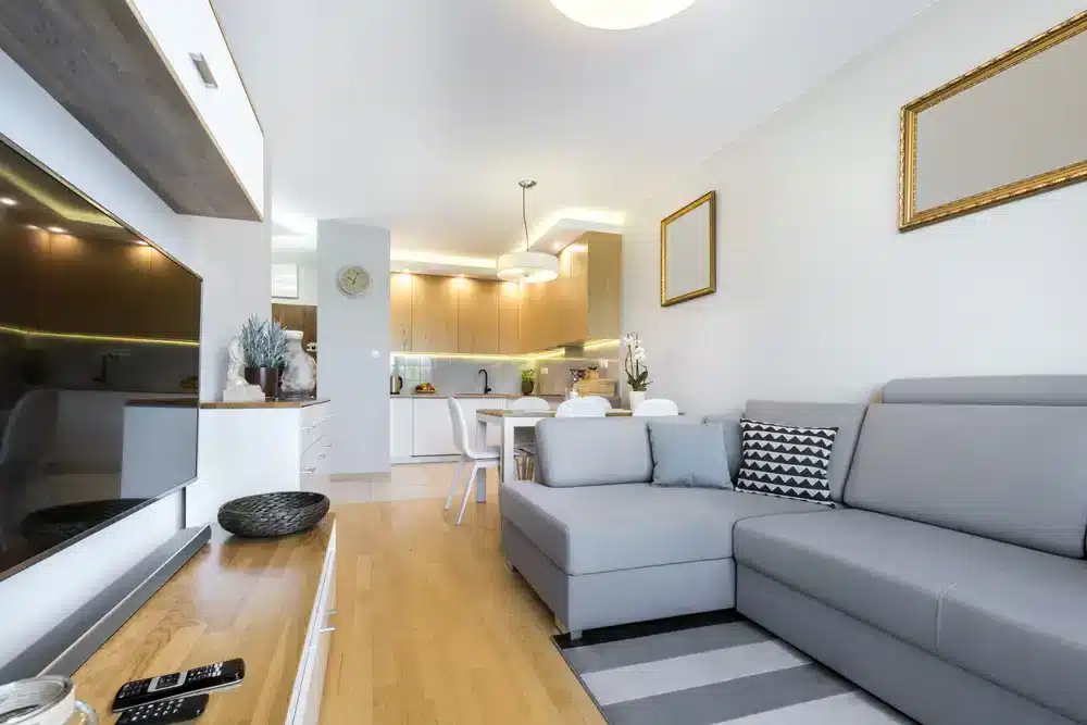 Svetlý obývací priestor s kuchynským kútom, biele skrinky a drevený stôl.