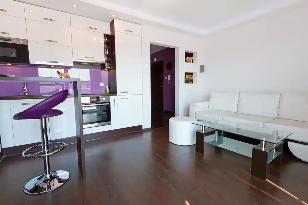 Moderný interiér garzónky s bielou kuchynskou linkou a fialovými prvkami. Centrálny kuchynský ostrovček s barovou stoličkou.