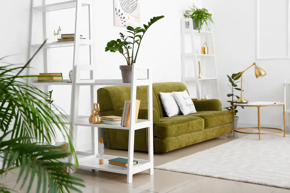 Obrázok ukazuje svetlú obývačku s výrazným zeleným zamatovým gaučom. Biele regály a zlaté doplnky pridávajú elegantný nádych, zatiaľ čo živé rastliny zdôrazňujú prírodné elementy.