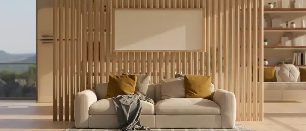 Na obrázku je moderná obývačka s drevenými lamelami, ktoré delia priestor a prinášajú dynamiku do interiéru. Pohovka v neutrálnych farbách s dekoratívnymi vankúšmi a prikrývkou dodáva pohodlie a štýl.
