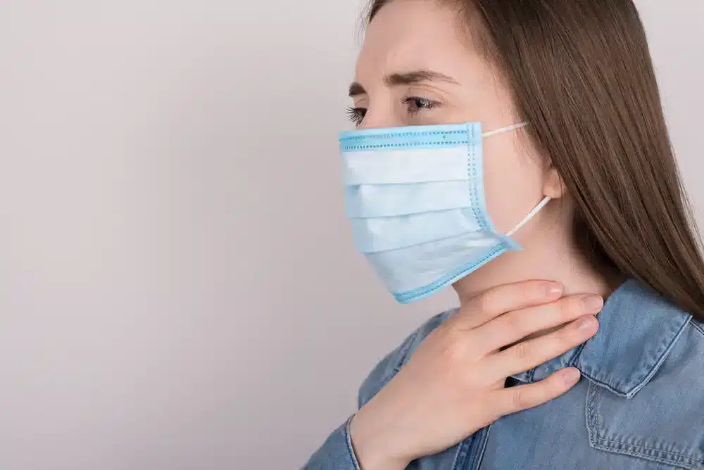 Žena s modrou ochrannou maskou na tvári sa dotýka hrdla, znak možnej bolesti hrdla alebo dýchacích ťažkostí.