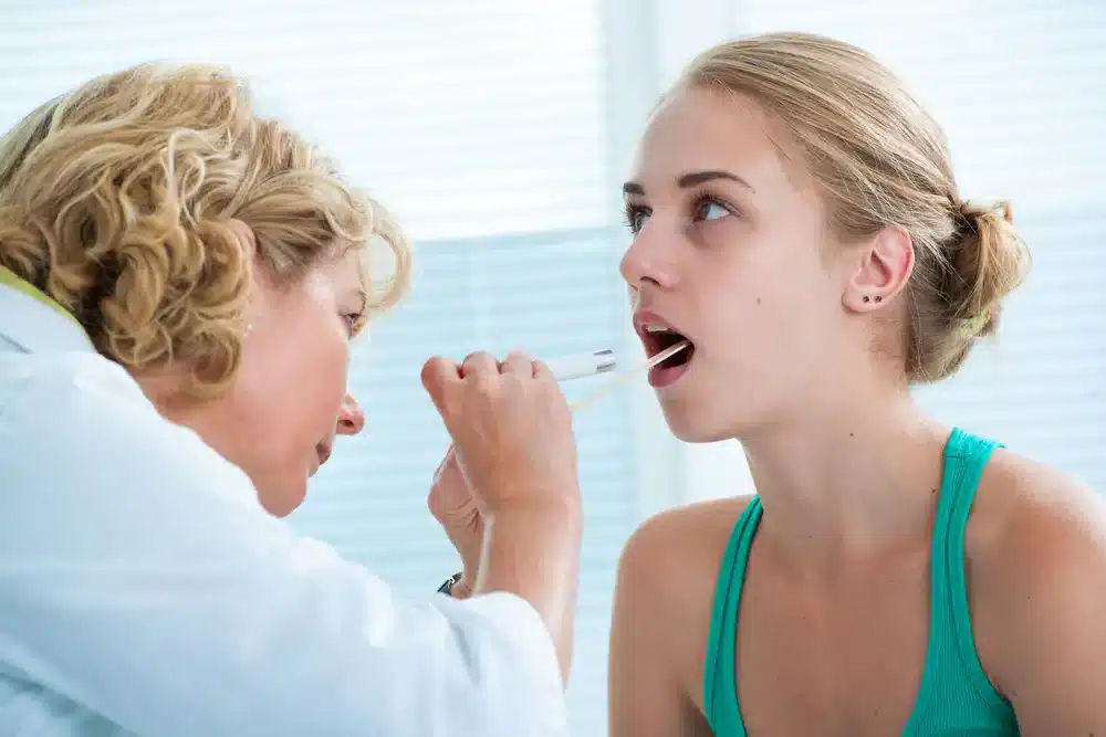Lekár vyšetruje hrdlo mladej ženy, čo je častý postup pri hľadaní príznakov zápalu priedušiek alebo iných respiračných ochorení.