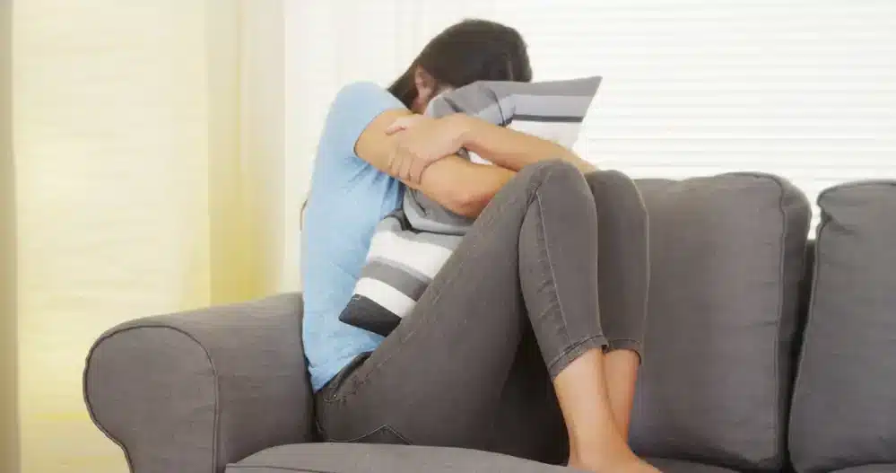 Žena objímajúca vankúš a zhrbená na gauči vyjadruje emočný a fyzický diskomfort počas menštruácie.