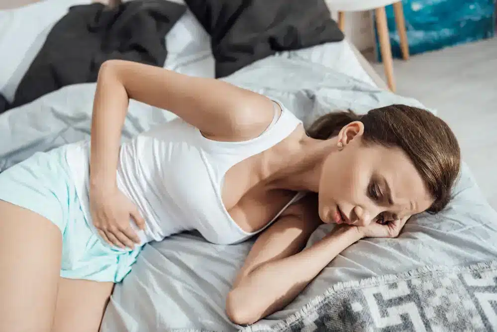 Žena ležiaca na boku v posteli s bolestivým výrazom v tvári a rukou na bruše symbolizuje problémy s menštruačným cyklom.