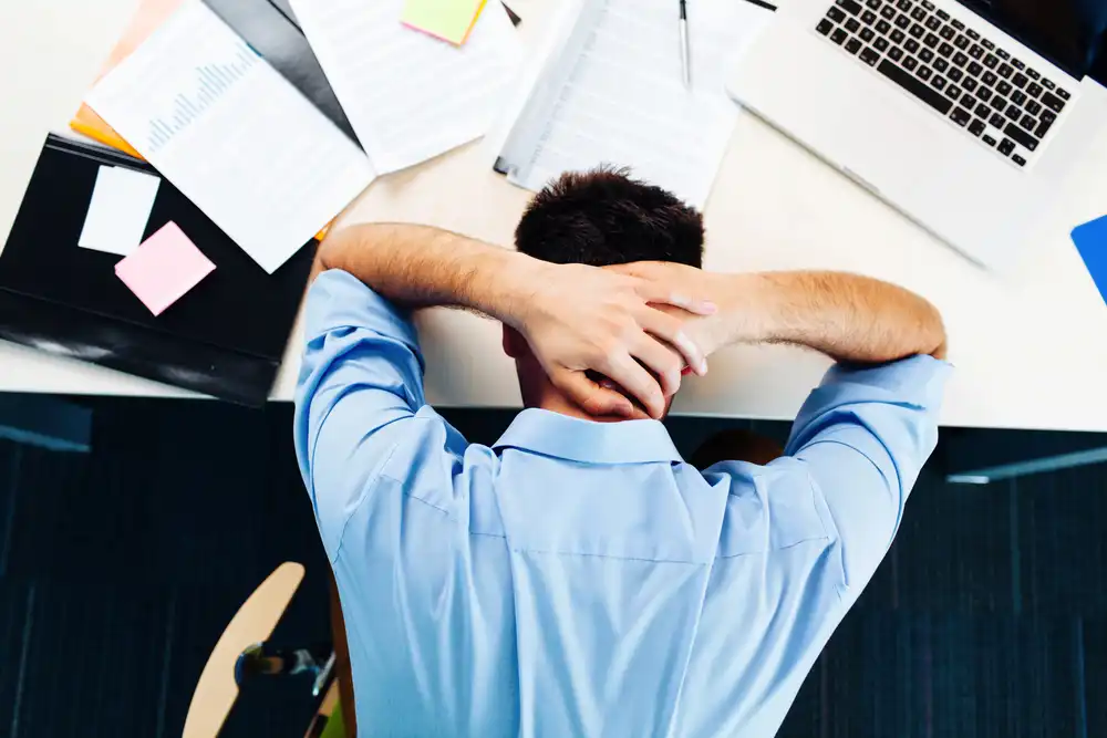 Muž v modrej košeli relaxuje v kancelárii s hlavou opretou do rúk, čo môže pomôcť znížiť stres a tým aj hladinu kortizolu.