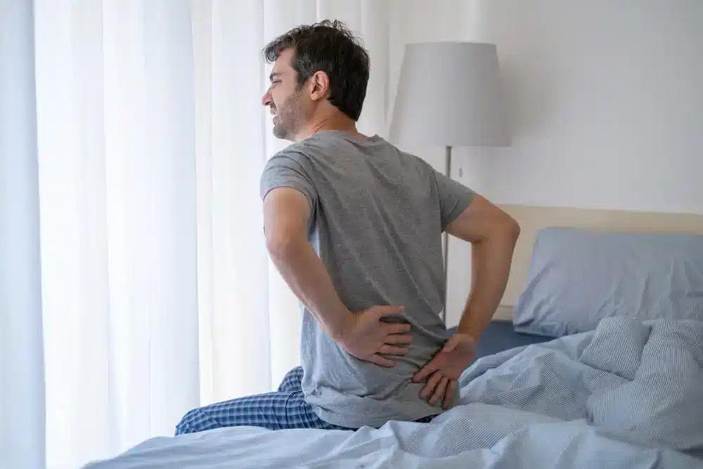 Muž sediaci na posteli a držiaci sa za bedrovú časť, vyjadruje bolesť, ktorá môže byť príznakom ischiadiky alebo zápalu sedacieho nervu.