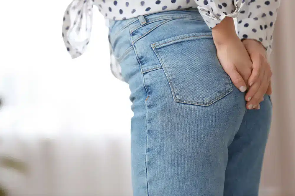 Žena v džínsoch si drží rukou zadok, čo môže symbolizovať diskomfort spôsobený hemoroidmi.