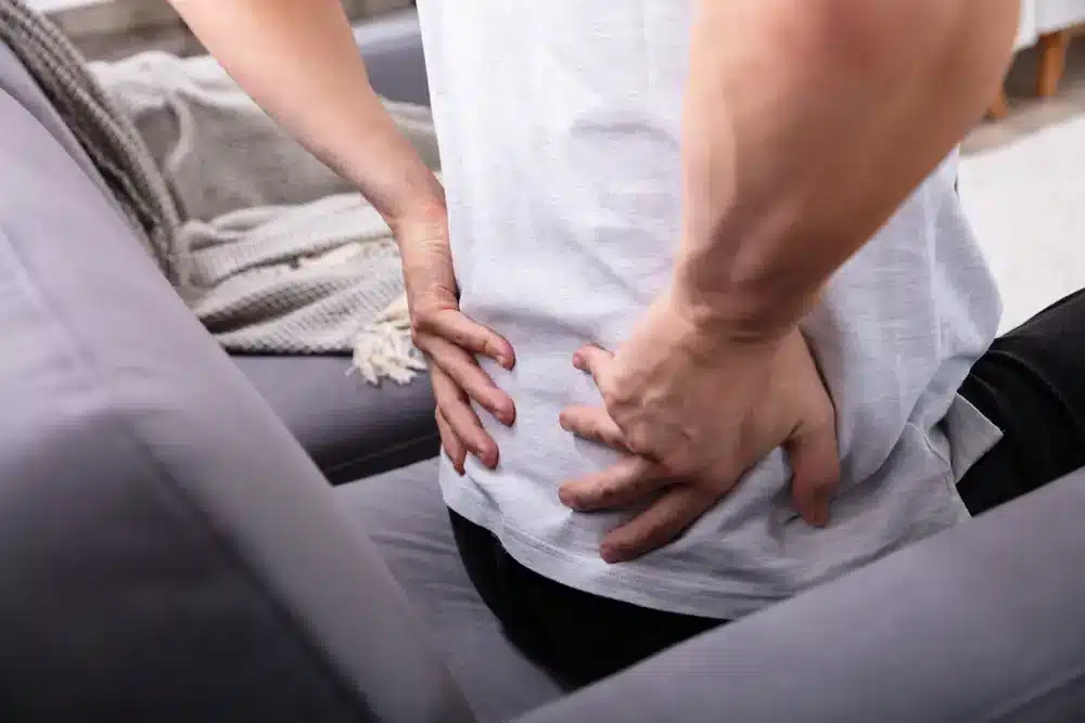 Muž sediaci na gauči a opatrne sa drží dolnej časti chrbta, čo môže ukazovať na ischias alebo zápal sedacieho nervu.