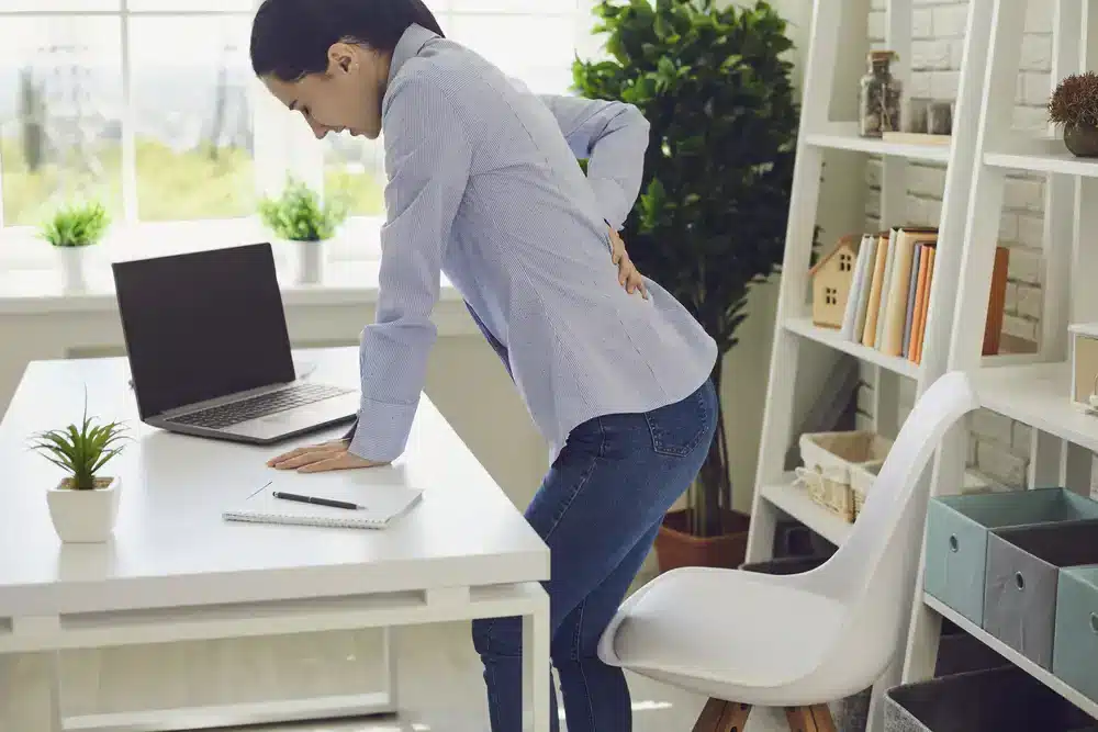Pracujúca žena v kancelárii sa predkláňa pred notebookom a drží sa za chrbát, čo naznačuje možný ischias alebo zápal sedacieho nervu.
