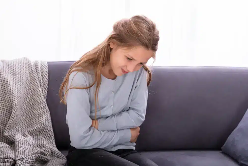 Mladé dievča sedí na pohovke, drží sa za brucho a máva bolesťou, čo naznačuje žalúdočné ťažkosti.
