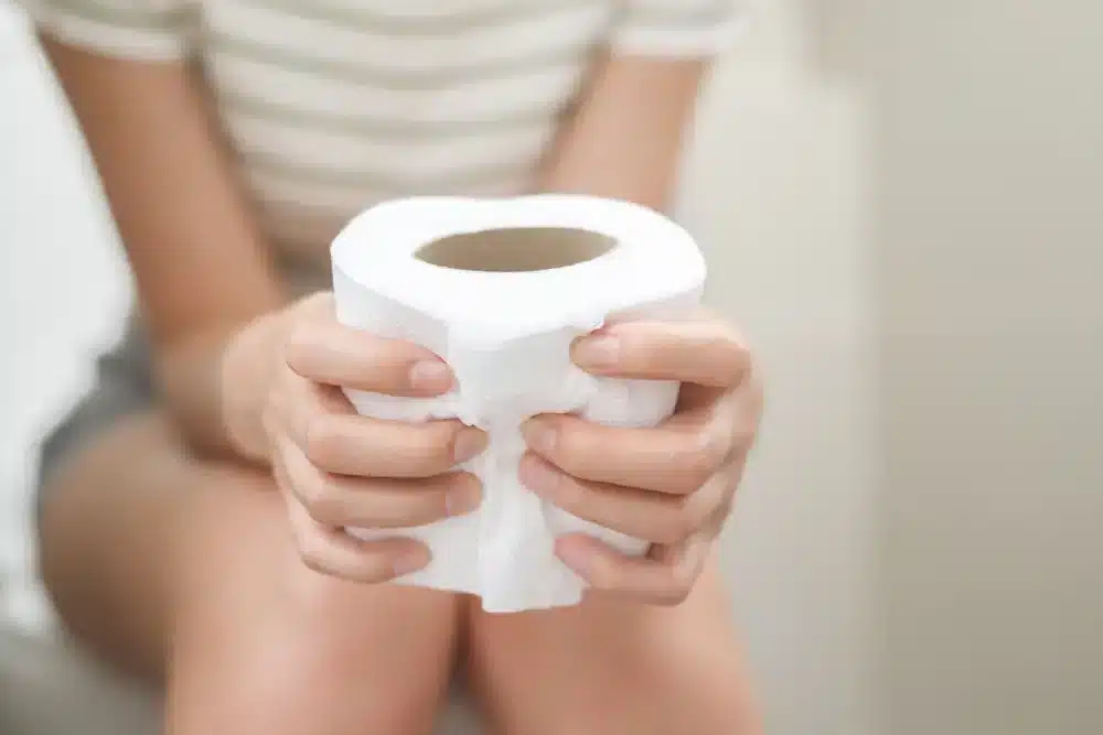 Žena sedí na toalete a drží toaletný papier.