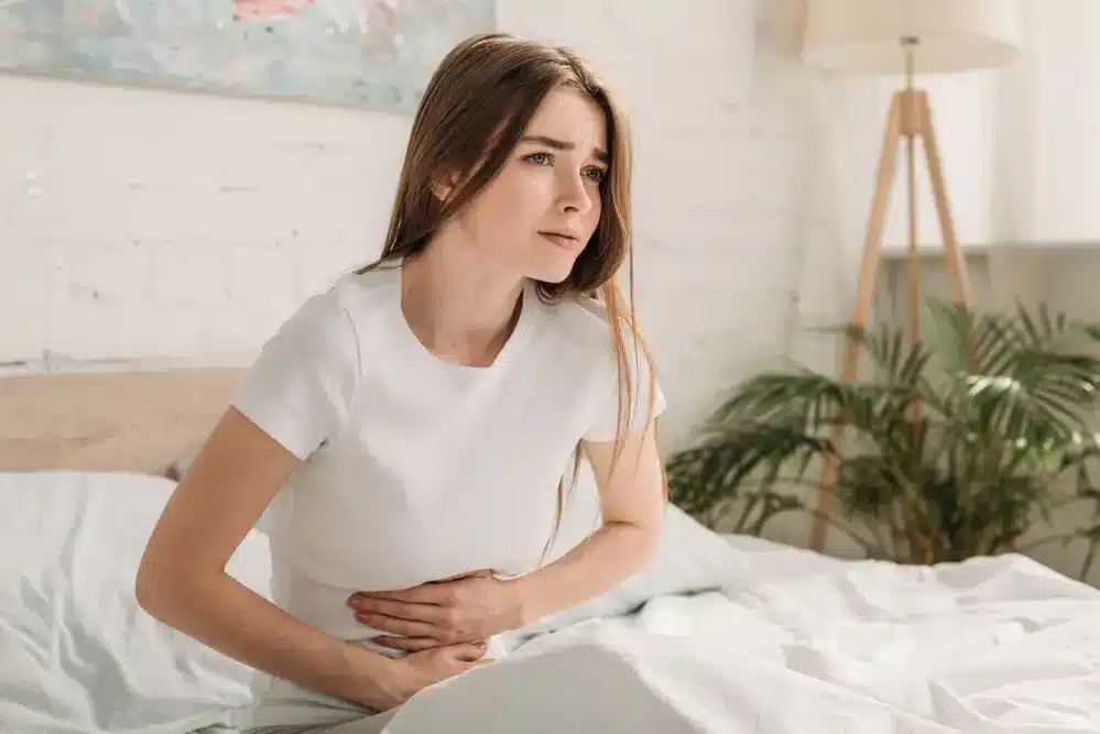 Mladá žena sedí na posteli s bolestivým výrazom a drží si brucho.